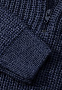 Tumši zilas krāsas adīta džemperis ar teksturētu rakstu, gludu rāvējslēdzēju un ribotiem piedurkņu galiem, kas piešķir papildu detaļas un piemērotību.