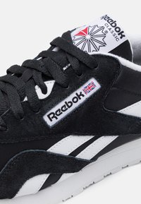 Zapatillas en blanco y negro con materiales de ante y textil; presentan un logo de Reebok, un acento de la bandera del Reino Unido y una suela texturizada.