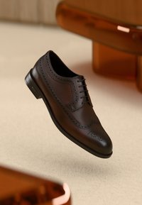 Zapato oxford de cuero marrón con detalles de brogue, diseño con cordones, punta redondeada y un acabado suave, sobre un fondo beige suave.