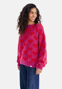 Růžový svetr s červeným srdíčkovým vzorem, kulatým výstřihem a volnými rukávy. Nosený s tmavými džínami. Měkká textura, ležérní design.
