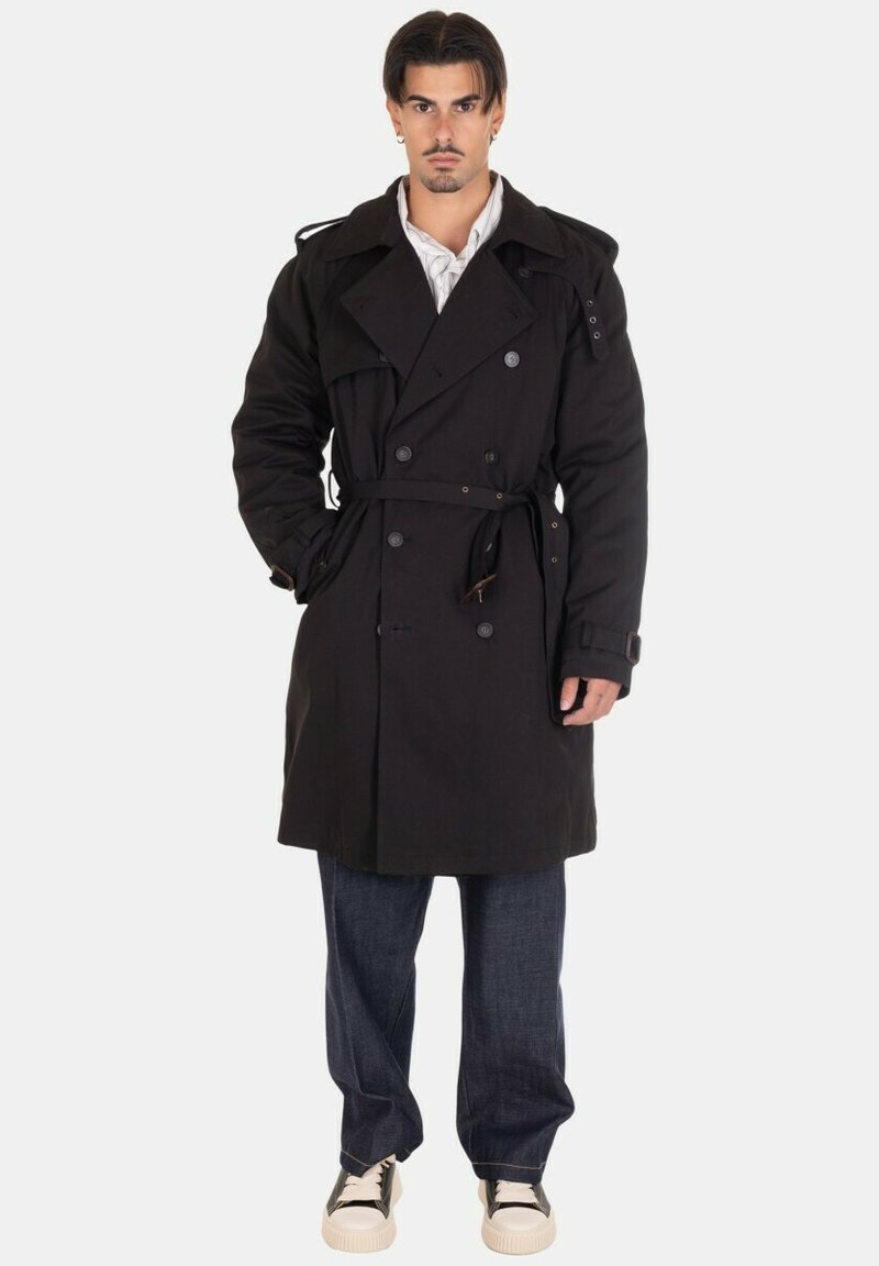 Impermeabile nero doppiopetto con vita legata, grandi revers e tasche laterali, indossato sopra jeans in denim scuri e sneaker.