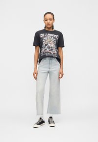 Νεαρή γυναίκα με πλεξουδάκια, φοράει μαύρο μπλουζάκι με τύπωμα, ανοιχτό μπλε φαρδιά τζιν, άσπρες κάλτσες και μαύρα αθλητικά παπούτσια, στέκεται σε λευκό φόντο.