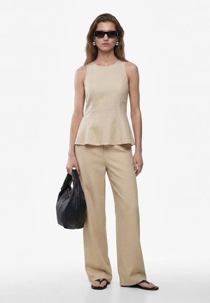 Vrouw draagt een beige mouwloos topje en broek, zwarte sandalen, zonnebril, grote zilveren oorbellen, houdt een zwarte leren handtas vast tegen een witte achtergrond.