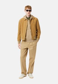 Cazadora bomber color beige, sudadera con capucha beige debajo, pantalones caqui, zapatillas blancas y gafas de sol negras. Tela suave, diseño minimalista y detalle de logotipo.