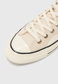 Beige canvas sneaker with white rubber toe cap and black trim. Features gold eyelets and cream laces, with textured upper and contrasting accents.