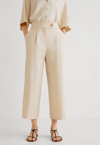 Beige wijde broek met plooien, hoge taille en een sluiting met een knop aan de voorkant. Gecombineerd met een bijpassend oversized overhemd met knopen en sandalen.