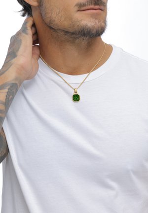 Homme portant un t-shirt blanc et un collier en or avec un pendentif octogonal vert, main touchant le cou, bras tatoué visible.