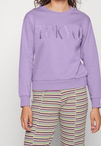Persona che indossa una felpa viola con "TOKYO" ricamato sul davanti e pantaloni a righe multicolori, in piedi contro uno sfondo semplice.