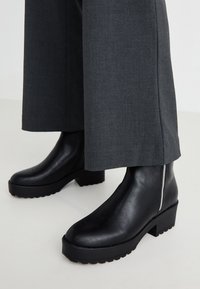 Botas de tobillo de cuero negro con un acabado texturizado, suela gruesa y cremallera lateral, llevadas con pantalones de pierna ancha grises.