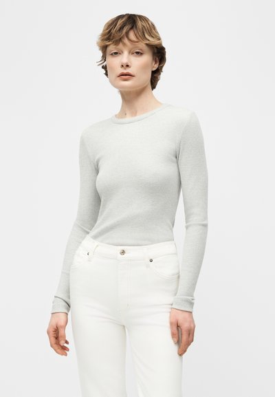 Γυναίκα με κοντά ανοιχτά καστανά μαλλιά που φοράει ανοιχτό γκρι ριγέ μακρυμάνικο τοπ και άσπρο παντελόνι ψηλόμεσο, σε απλό φόντο.