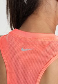 Camiseta sin mangas de Nike en tejido de malla naranja neón con cuello redondo, que presenta un logo plateado en la parte posterior y un patrón texturizado.