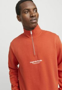 Ruskofärgad zip-up sweatshirt med hög krage, texturerat tyg och vit "Originals Studio"-logotyp på framsidan.