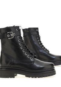 Paire de bottes en cuir noir à lacets avec semelles épaisses et une sangle à boucle autour de la cheville, présentées sur un fond blanc.