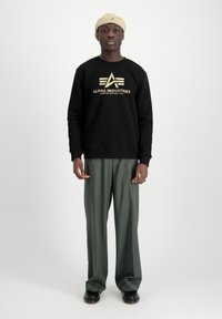 Fekete kapucnis pulóver arany "ALPHA INDUSTRIES" logóval, zöld, szabott nadrággal és bézs kötött sapkával párosítva.