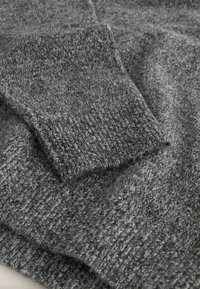 Harmaa neulottu villapaitakangas, jossa on teksturoitu pinta ja taitettu kalvosinn yksityiskohtana. Pehmeä, keskipainoinen materiaali näkyvillä saumoilla.