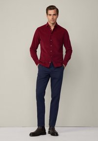 Camicia button-up color marrone scuro con una texture liscia, abbinata a pantaloni navy su misura e stivali neri alla caviglia. Design semplice, silhouette aderente.