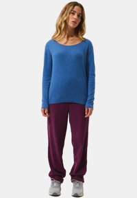 Blauer Wollpullover mit rundem Ausschnitt, kombiniert mit lila gerippten Hosen. Das Modell trägt graue Sportschuhe und zeigt einen lässigen, entspannten Look.