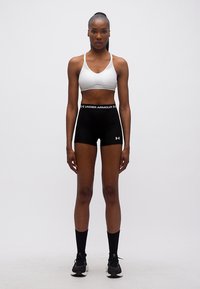 Αθλητική γυναίκα στέκεται μπροστά, φοράει λευκό αθλητικό σουτιέν, μαύρο σορτς Under Armour, μαύρες κάλτσες και μαύρα αθλητικά παπούτσια.