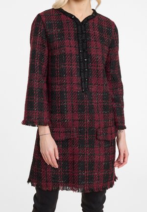 Femme portant une robe texturée à carreaux bordeaux et noire, longueur genou, avec des manches trois-quarts et des détails à franges aux poignets et à l'ourlet.