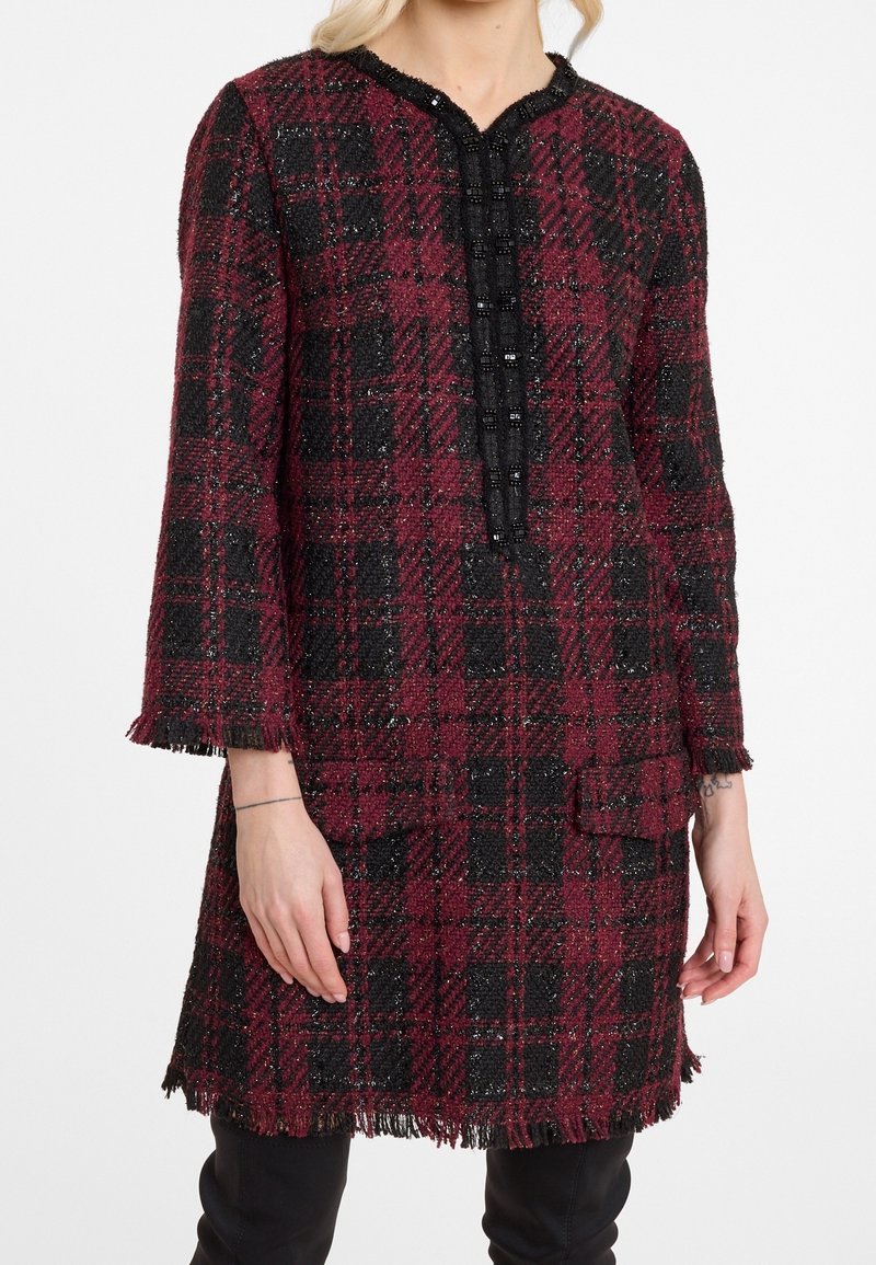 Femme portant une robe texturée à carreaux bordeaux et noire, longueur genou, avec des manches trois-quarts et des détails à franges aux poignets et à l'ourlet.