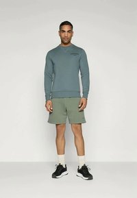 Harmaa huppari, jossa on logo rinnassa, yhdistettynä vaaleanvihreisiin shortseihin. Mustat urheilukengät, valkoiset sukat ja tasainen tausta.