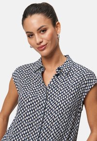 Blusa a righe navy e crema con colletto, maniche a sbuffo e chiusura frontale con bottoni. Tessuto leggero e testurizzato con design geometrico.