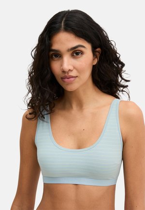 Young woman with curly dark hair wearing a light blue striped sleeveless crop top against a plain background.
