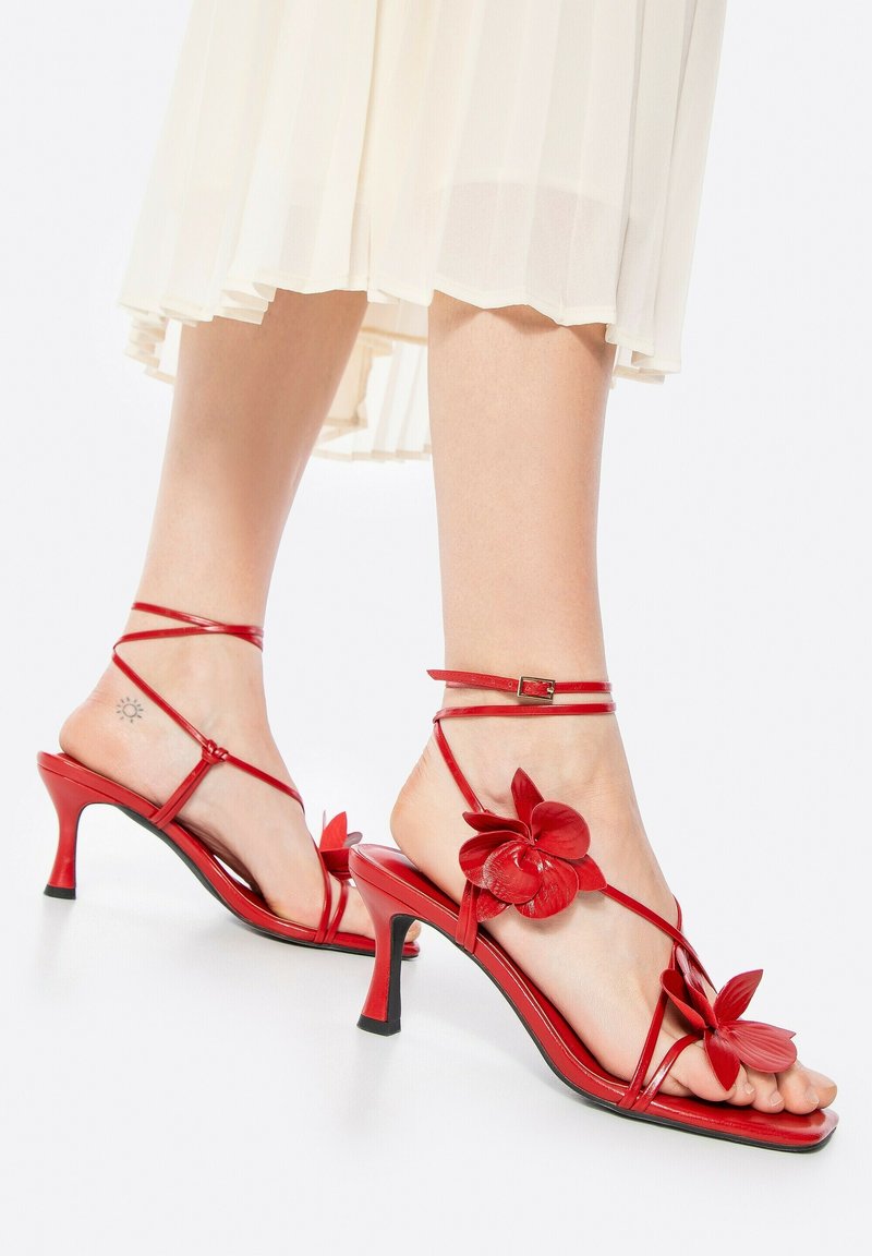 Rote High Heels mit einem floralen Design, dünnen Riemen und einer eleganten, glänzenden Oberfläche. Der Absatz ist etwa 8 cm hoch.