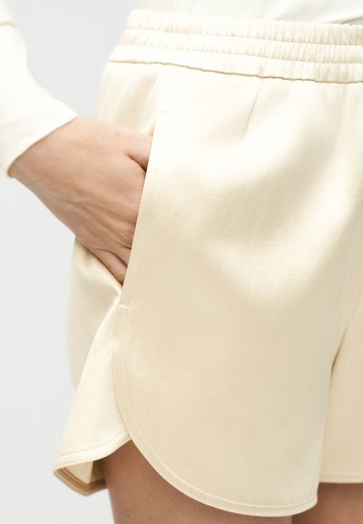 Χέρι στην τσέπη ενός μπεζ σορτς με ελαστική μέση και στρογγυλεμένη ραφή στο κάτω μέρος, φορεμένο με μακρυμάνικη μπλούζα ίδιου χρώματος.