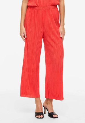 Femme portant un pantalon corail plissé à jambes larges et taille haute, assorti à des sandales noires à talons ouverts, sur un fond uni.