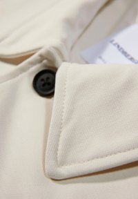 Ljust beige jacktyg med en strukturerad yta, som har en svart knapp och en vikkrage. En skötselrådsetikett är delvis synlig.