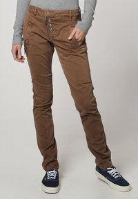 Brown slim-fit trousers made of cotton blend, featuring a button-up fly, side pockets, and a smooth texture. Paired with navy sneakers.