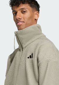 Fleece zip-up jacket in light green with high collar. Features a textured surface and black Adidas logo on the chest. Zipper pull accent included.