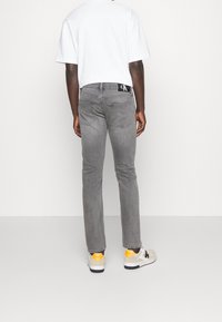 Man som bär grå jeans, vit T-shirt och beige sneakers med orangea detaljer, står vänd mot en vit vägg.