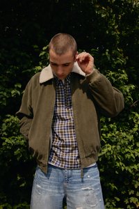 Chaqueta bomber de pana verde oliva con un cuello de forro polar beige, superpuesta sobre una camisa a cuadros, combinada con jeans azul desgastados.