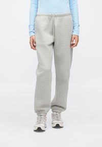 Άτομο που φοράει ανοιχτό γκρι φόρμες με ελαστικούς ιμάντες, ανοιχτό μπλε ριγέ μακρυμάνικο μπλουζάκι και λευκά αθλητικά παπούτσια, που στέκεται σε άσπρο φόντο.