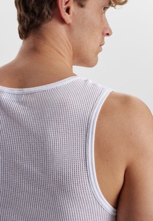 Hvid mesh tanktop med rund hals og brede ærmegab, der har et tekstureret mønster og er lavet af bomuldsmateriale. Bagudsigten viser en tætsiddende pasform.