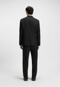 Mees seisab seljaga vaataja poole, kandes musta ülikonda koos sobivate pükste ja mustade kingadega heleda ühevärvilise tausta ees.