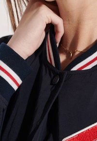 Marinblå bombarjacka med en röd och vit randig ribbad krage och muddar, samt rund knappstängning och en strukturerad tyg. Guldhalsband synligt.