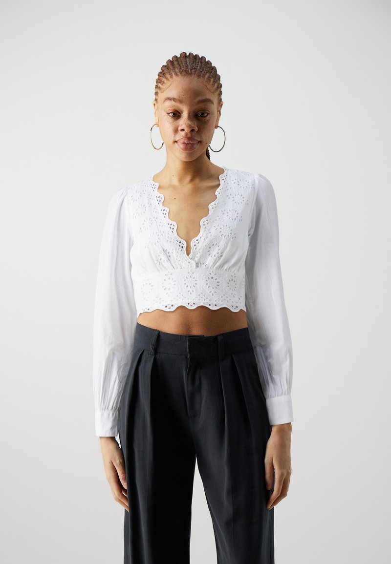 White cropped blouse with long, puffed sleeves, lace detailing along the neckline, and a scalloped hem. Paired with dark, pleated trousers.