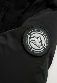 Sort jakke materiale med en cirkulær, tekstureret patch med et bjergdesign og teksten "Clarks" i midten, omkranset af sort.