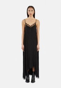 Fekete maxi ruha csipkével díszített V-nyakkivágással, vékony spagettipántokkal és magas-alacsony szegéllyel; fekete bokacsizmával párosítva.