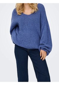 Femme portant un pull en maille bleu ample avec des manches ballon et un jean bleu foncé, debout devant un fond blanc uni.