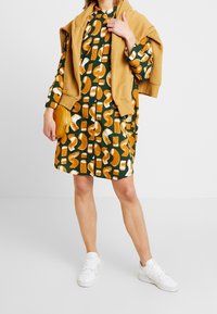 Grön mönstrad skjortklänning med stora orange och vita geometriska mönster, med lång ärm uppvikt och en beige tröja draperad över axlarna.