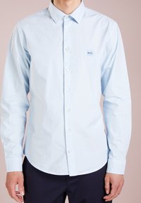 Světle modrá košile s knoflíky z bavlny. Má klasický límec, dlouhé rukávy a malou značkovou nášivku na levé straně hrudi.