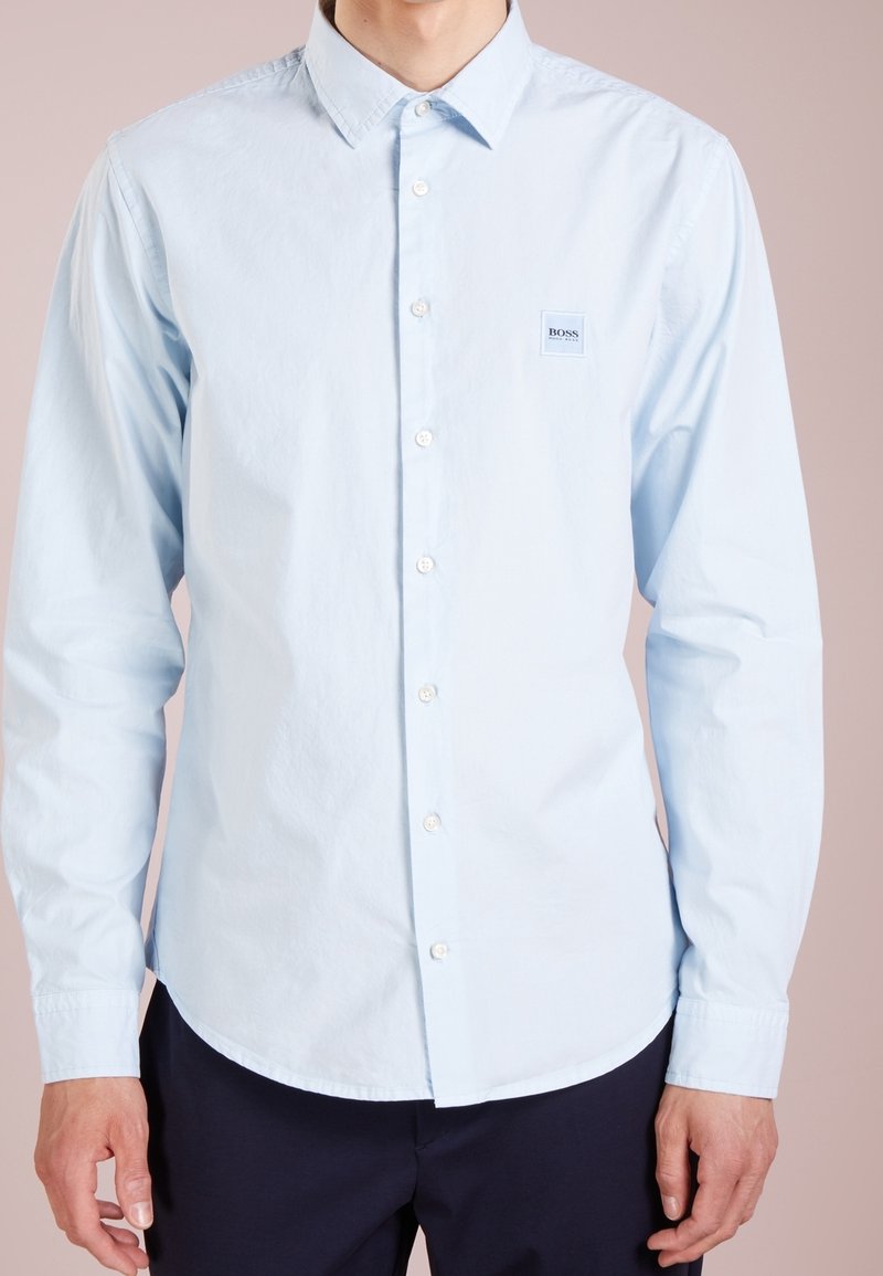 Světle modrá košile s knoflíky z bavlny. Má klasický límec, dlouhé rukávy a malou značkovou nášivku na levé straně hrudi.