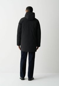 Juodas vandeniui atsparus parka su gobtuvu, pasižymintis lygia tekstūra, ilgomis rankovėmis ir iki kelių siekiančiu dizainu. Dėvimas su tamsiai mėlynais džinsais ir juodais batais.