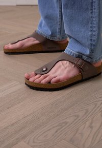 Ruske flip-flop sandale s teksturiranim đonom, detaljem kopče i plutenim uloškom, nosene s plavim trapericama, postavljene na drveni pod.