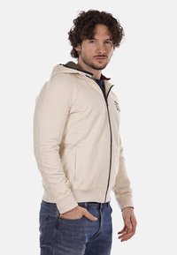 Felpa beige con zip e cappuccio, tasche laterali, polsini e fondo a coste, con colletto scuro a contrasto e logo sul petto.