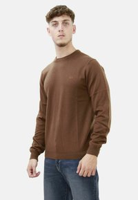 Maglione marrone a maniche lunghe in un tessuto a maglia liscia, con polsini e orlo a costine, e un piccolo logo arancione sul lato sinistro del petto.